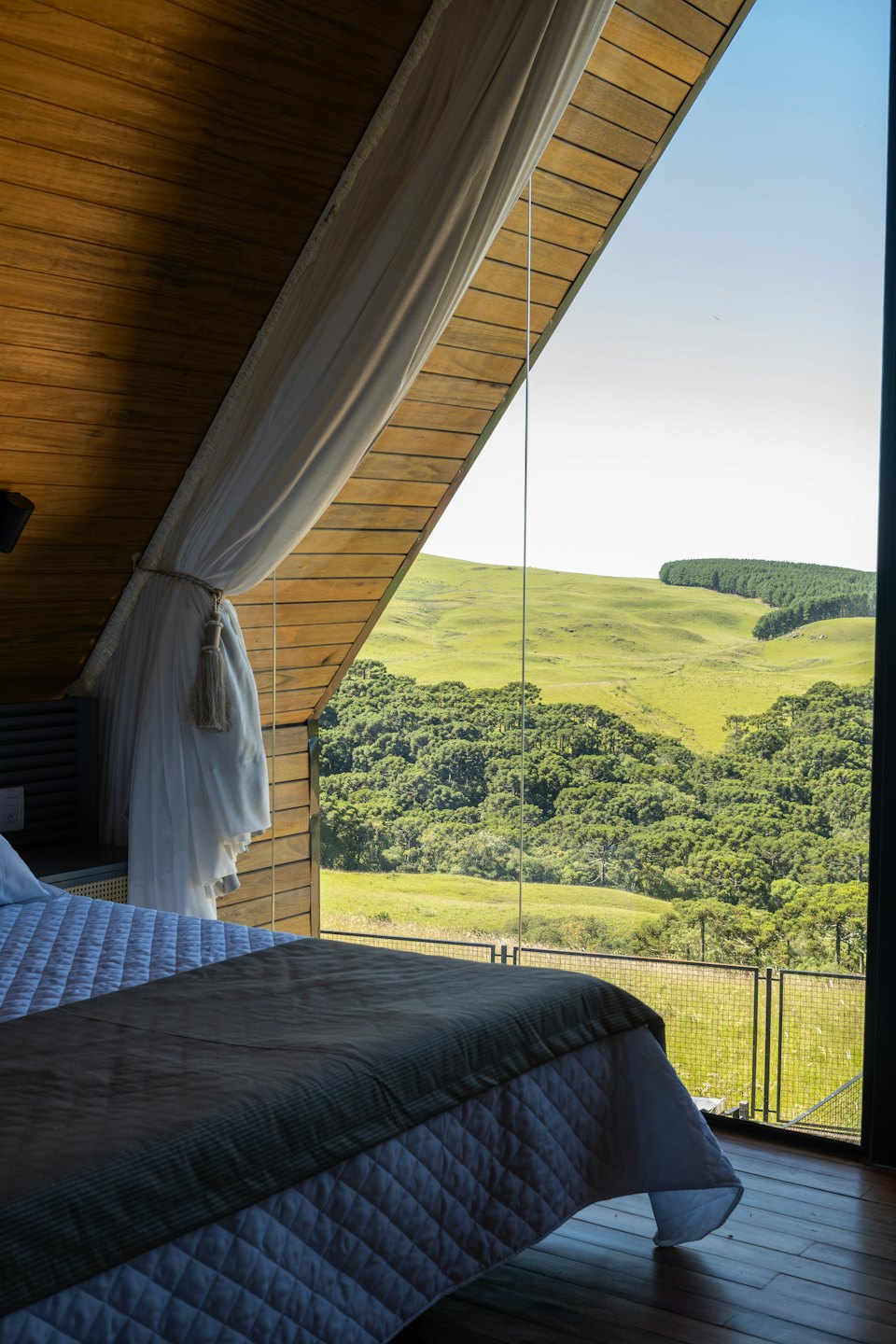 Camera da letto con grande finestra triangolare che si affaccia su colline verdi e foresta.